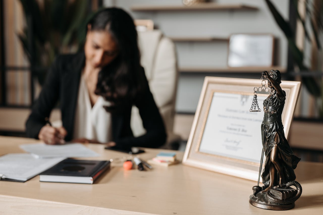 why-choose-us Focused woman writing at desk with Lady Justice figurine and certificate in office.